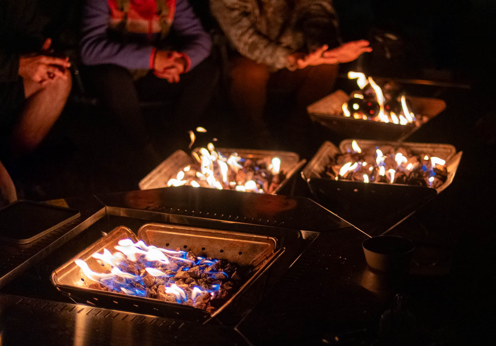 Night scene with four Snow Peak Takibi fire pits powered by Zutto SP1 smokeless propane gas adapters as people gather and warm their hands around the flames