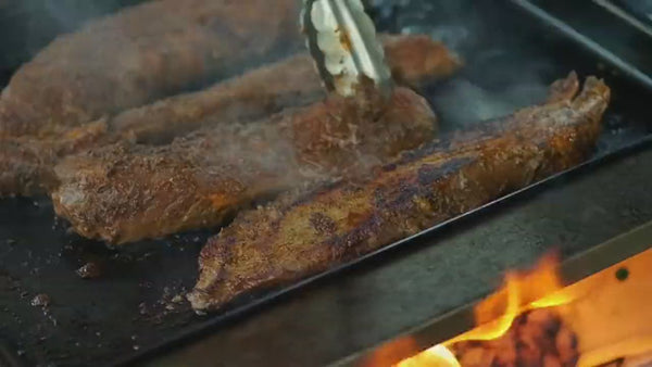Slow motion video of marinated beef sizzling and being flipped on a Snow Peak griddle heated by the Zutto SP1 smokeless propane gas adapter installed in a Snow Peak Takibi fire pit